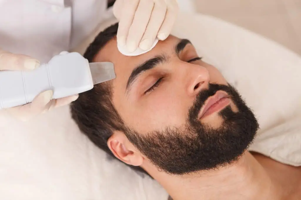A man with a beard lies with his eyes closed while a person wearing gloves uses a cotton wool pad and a handheld skincare device on his forehead, possibly for a facial treatment.