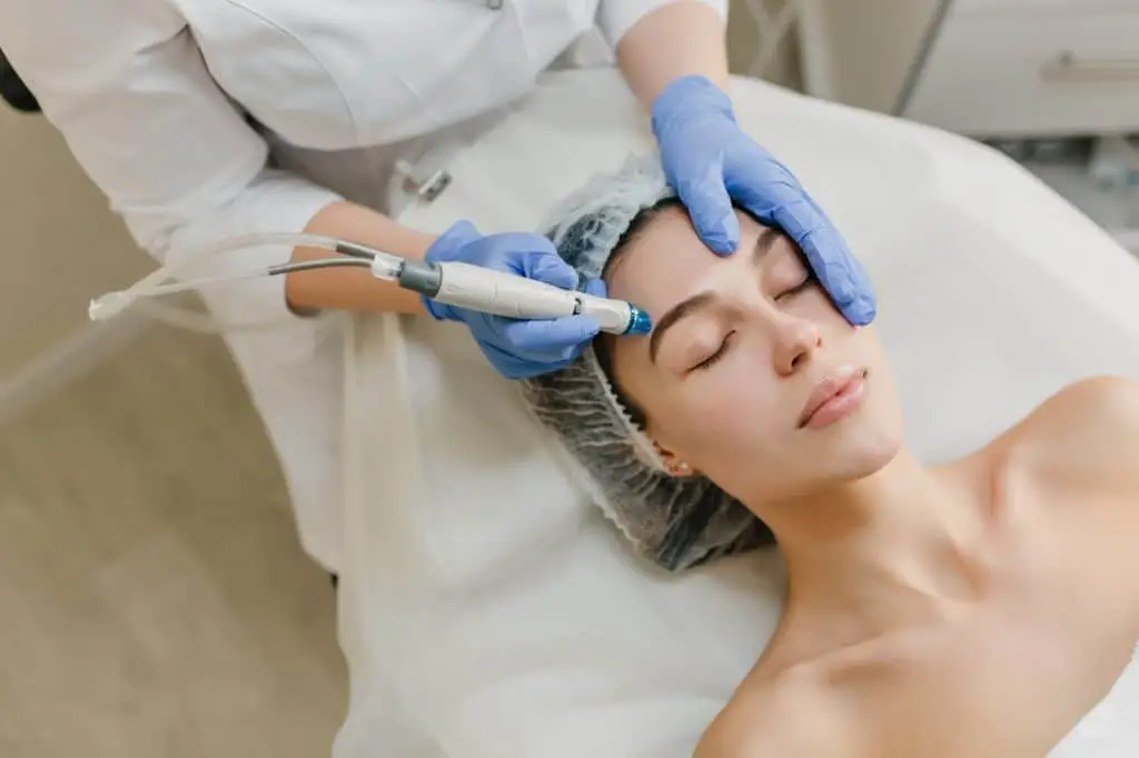 A person lies with eyes closed while a professional wearing gloves uses a skincare device on their face. The person’s hair is covered with a disposable cap, and the setting appears to be a clinic or spa.